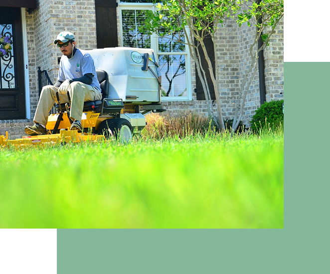 A person operating a riding lawn mower to cut grass in front of a brick house as part of residential landscaping.